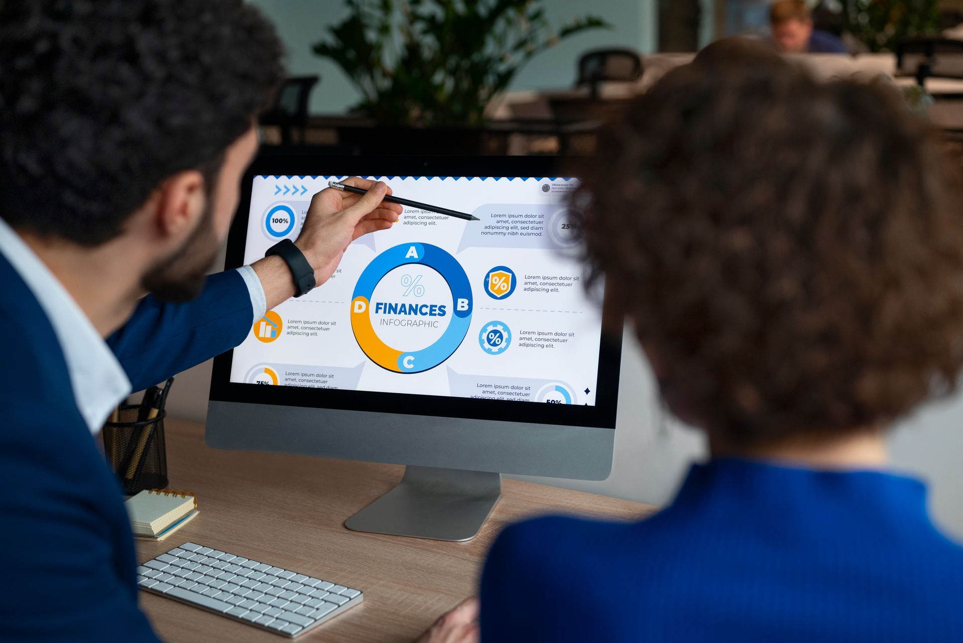 Man points at a circular chart on a computer screen while explaining to a woman at a desk in an office.