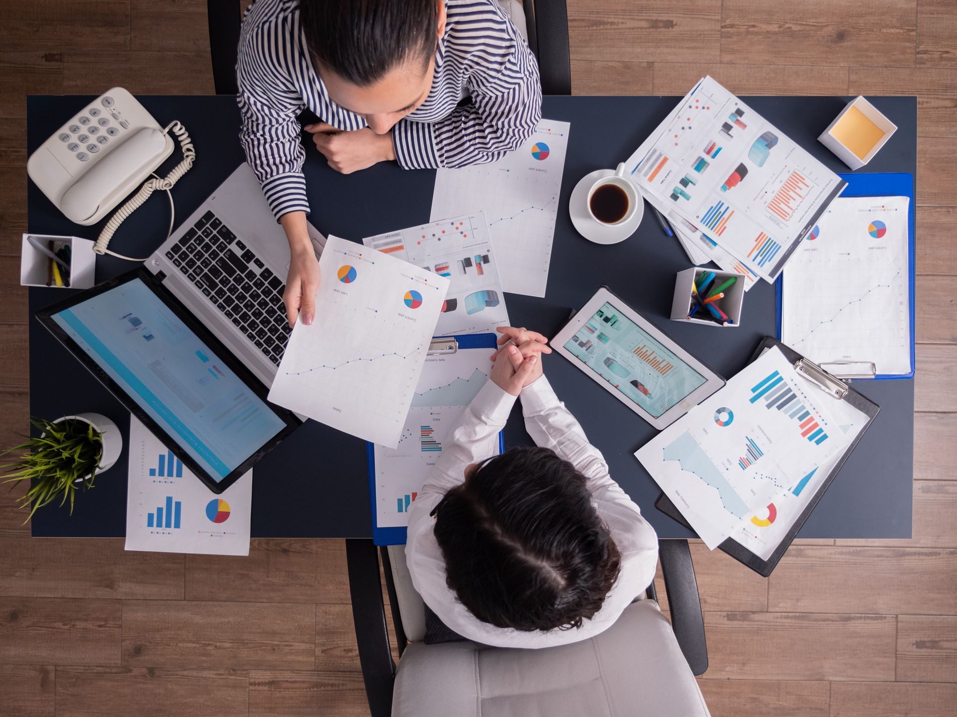 Two people at a desk reviewing data reports, laptop, tablet, and coffee present.