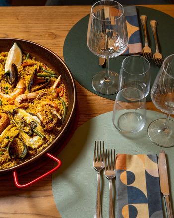 Paella in padella su un tavolo apparecchiato con bicchieri da vino, tovagliette e posate.
