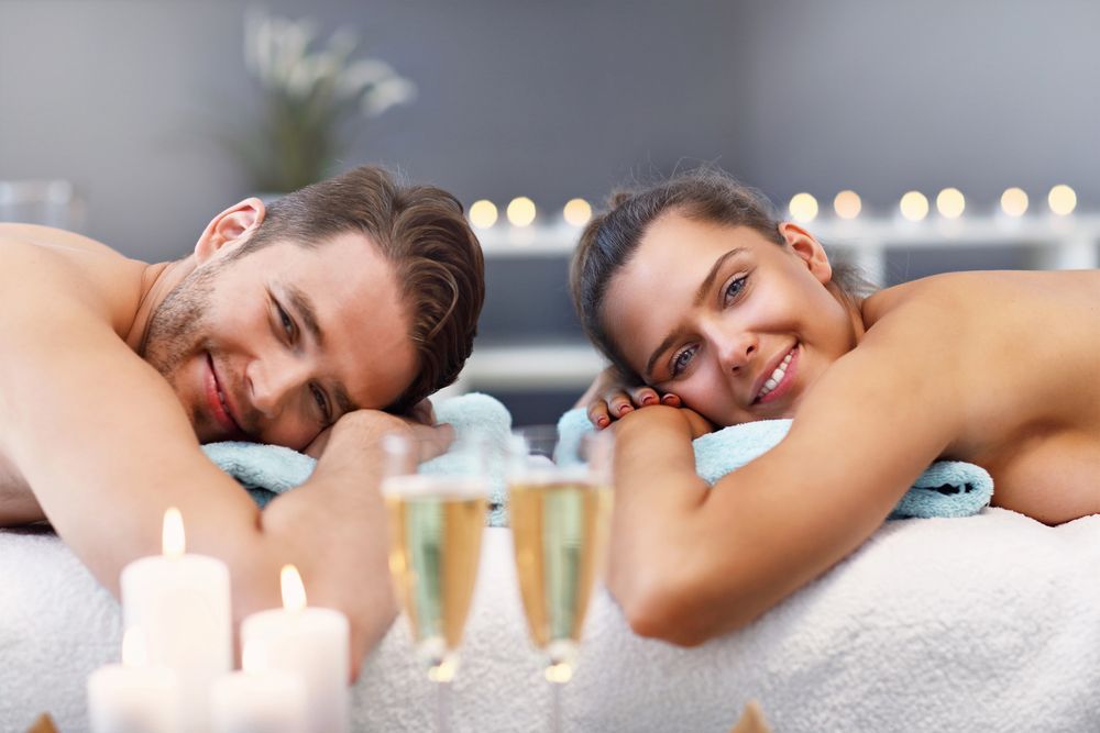 Due persone si rilassano in una spa, appoggiate ad asciugamani, con candele accese in primo piano.
