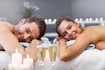 Due persone si rilassano in una spa, appoggiate ad asciugamani, con candele accese in primo piano.