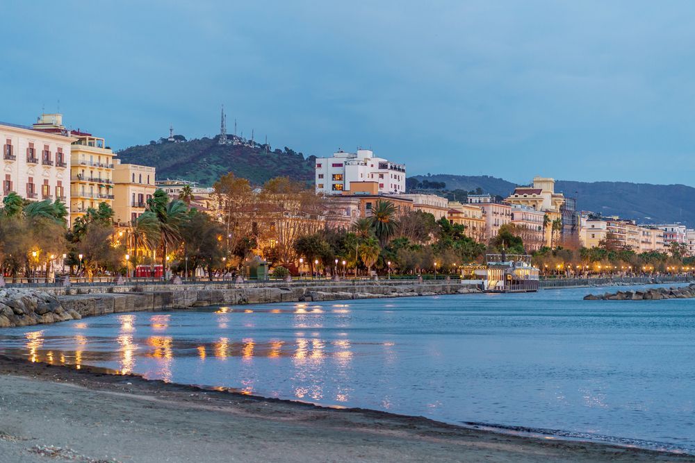 Tramonto su una passeggiata costiera con edifici illuminati, palme e un mare calmo che riflette le luci