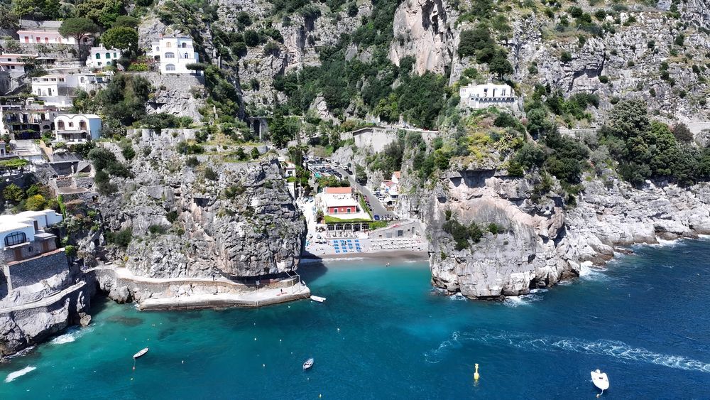 Insenatura turchese con scogliere, case bianche, una piccola spiaggia sabbiosa e barche su acque cristalline.