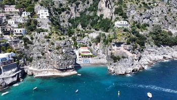 Villaggio costiero a picco sul mare, con case bianche e una piccola caletta turchese ai piedi di colline rocciose.