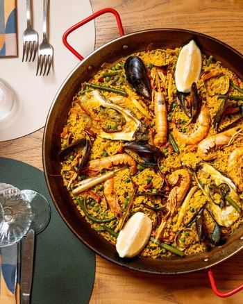 Paella di pesce in una padella rossa su un tavolo di legno, con bicchieri da vino e posate nelle vicinanze.