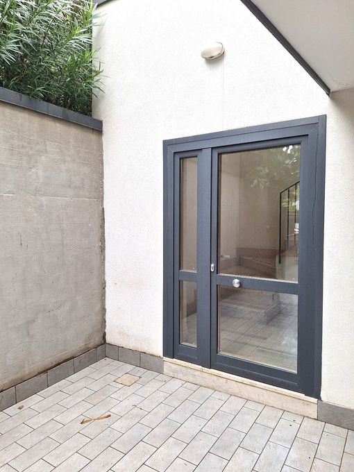 Piccolo patio con pavimentazione grigio chiaro, parete bianca e porta a vetri grigio scuro che conduce all'interno.