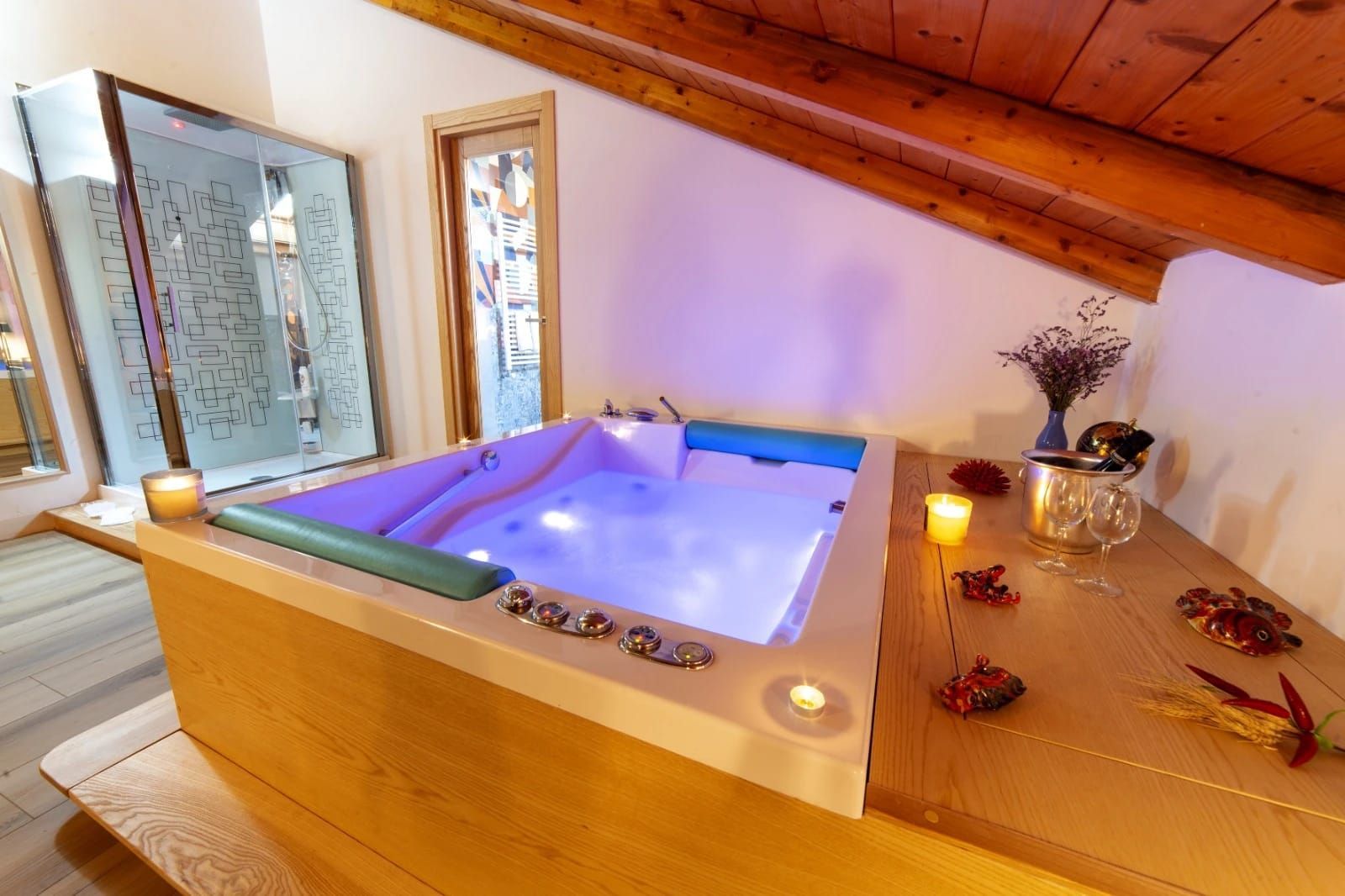 Accogliente sala spa in mansarda con vasca idromassaggio illuminata, soffitto rivestito in legno e candele su una mensola di legno.