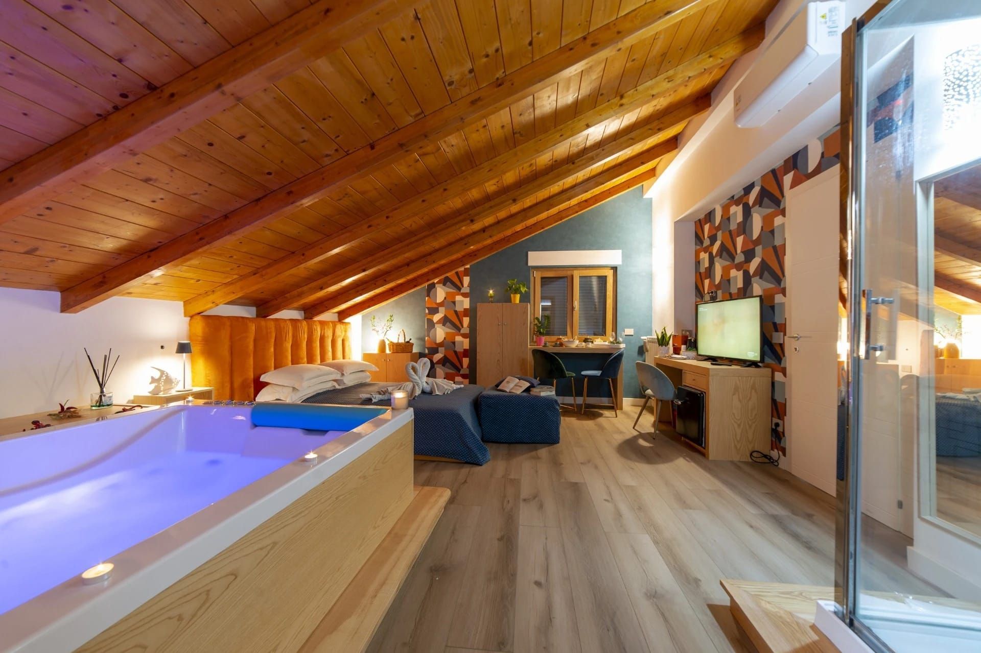 Camera da letto mansardata moderna con soffitto in legno inclinato, letto king size, poltrone e vasca da bagno illuminata di blu.