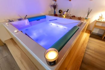 Vasca idromassaggio interna illuminata con acqua blu, candele agli angoli e una terrazza in legno accanto.