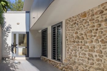 Moderno passaggio pedonale nel cortile adiacente a una casa in pietra e bianca con porte e finestre in vetro.
