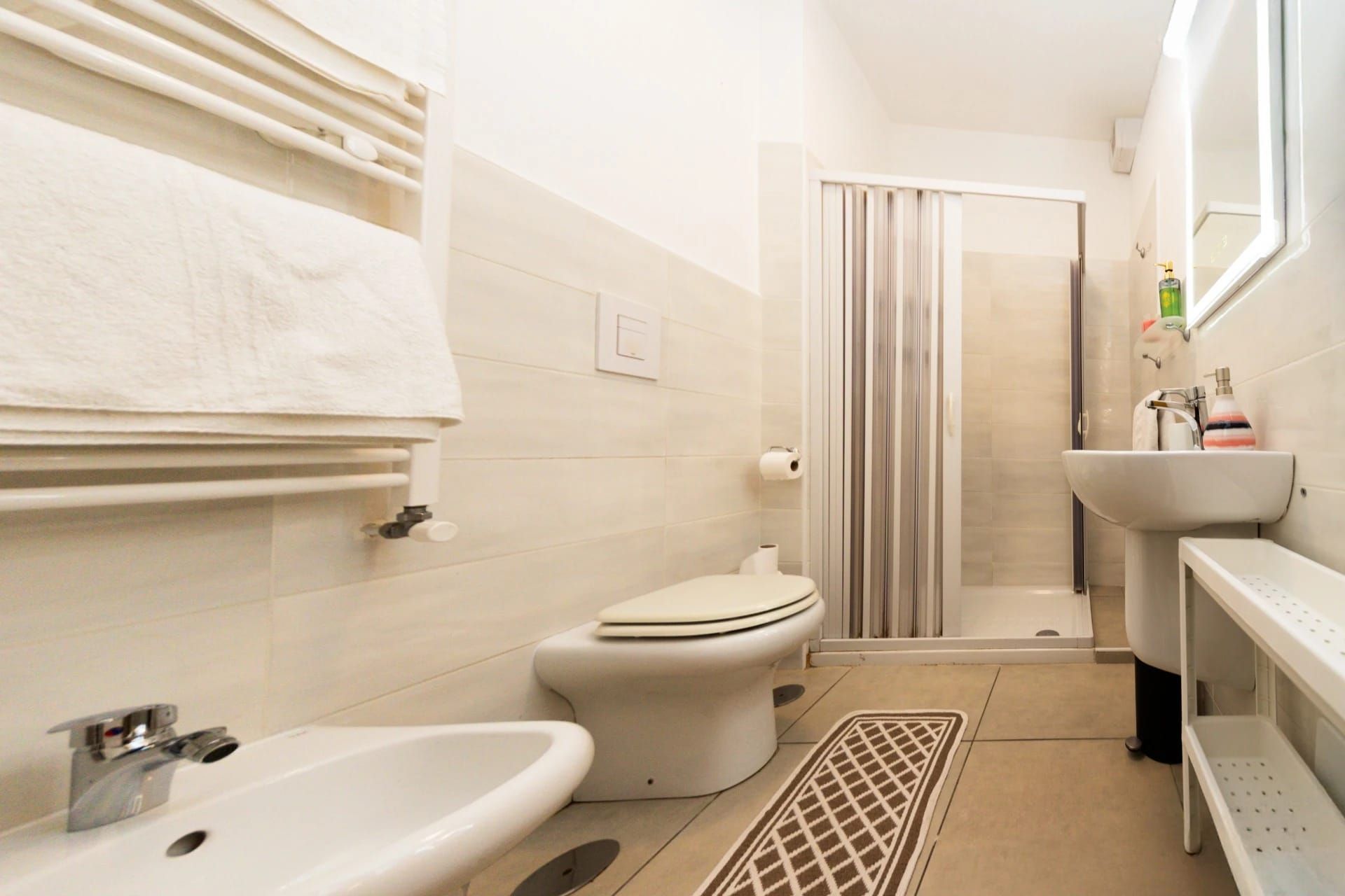 Bagno luminoso color beige con WC, bidet, lavabo, portasciugamani e box doccia in vetro.