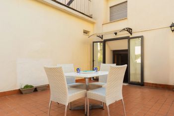 Patio con tavolo rotondo e sedie in vimini all'esterno di un edificio color crema con porte a vetri aperte.