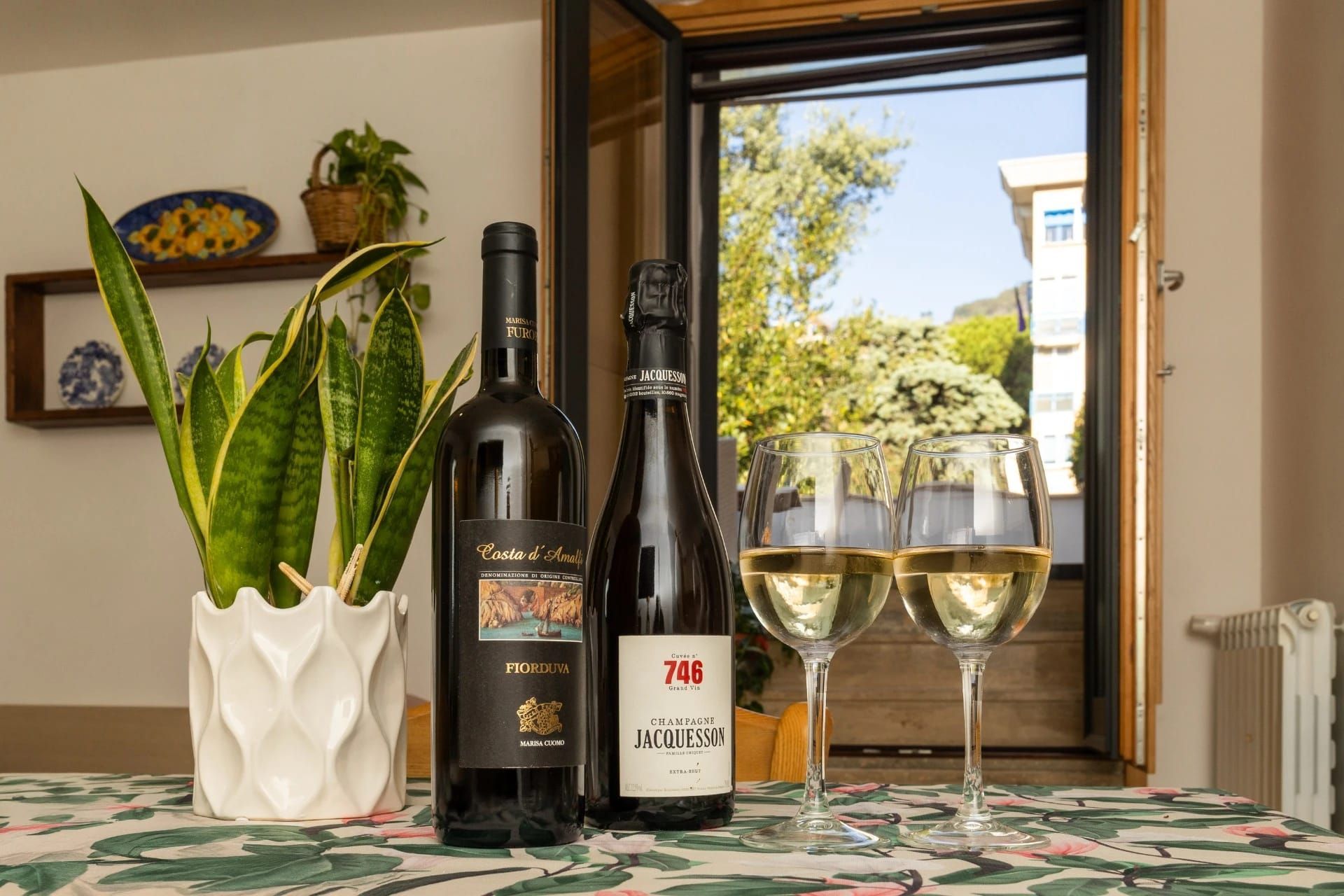 Un tavolo con bottiglie di vino, due bicchieri e una pianta di sansevieria in vaso accanto a una finestra aperta.