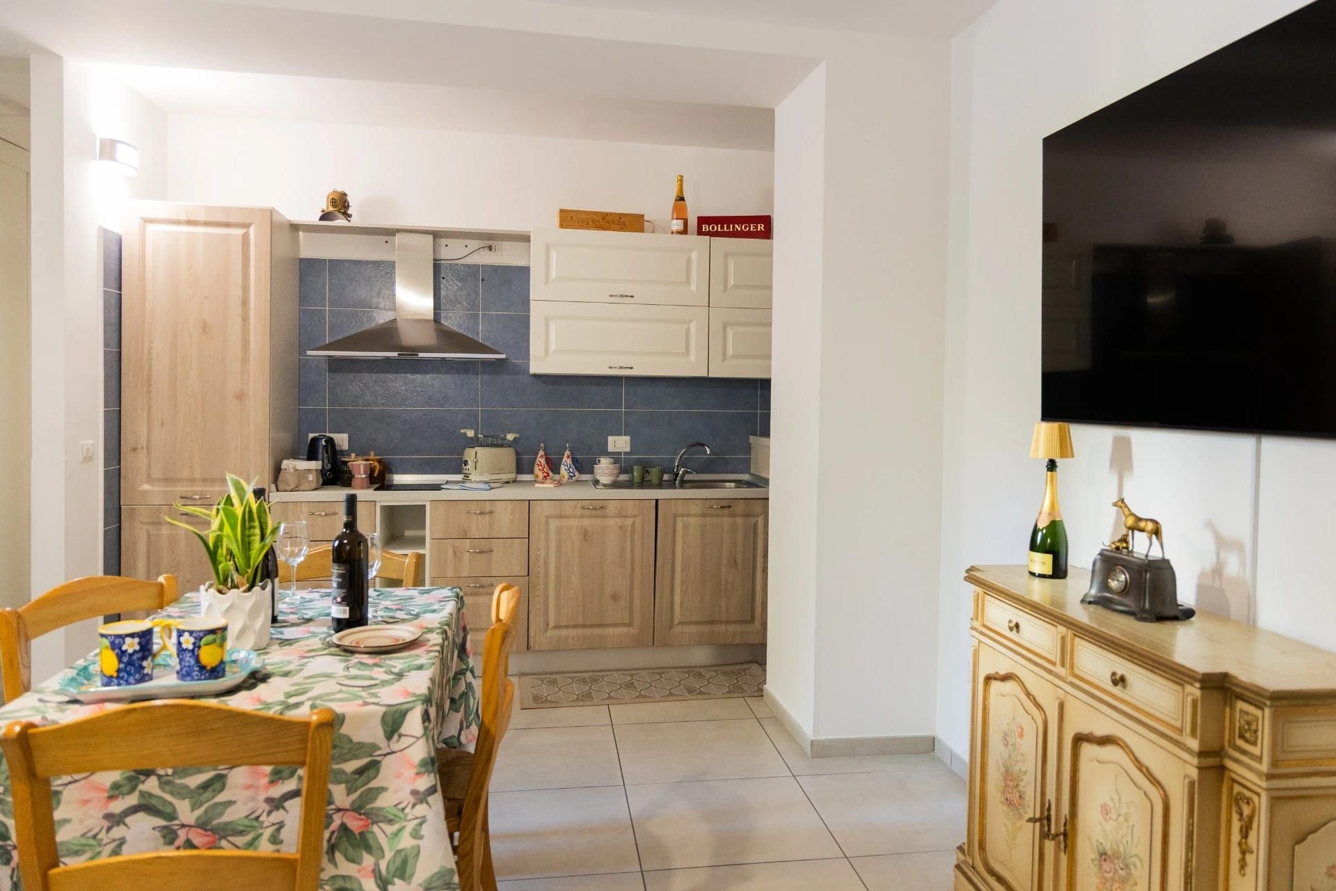 Zona pranzo che si apre su una cucina con mobili in legno, un tavolo apparecchiato con stoviglie e un televisore a parete.