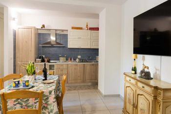Zona pranzo con tavolo apparecchiato che si apre su una cucina in legno chiaro e un televisore a parete sulla destra.