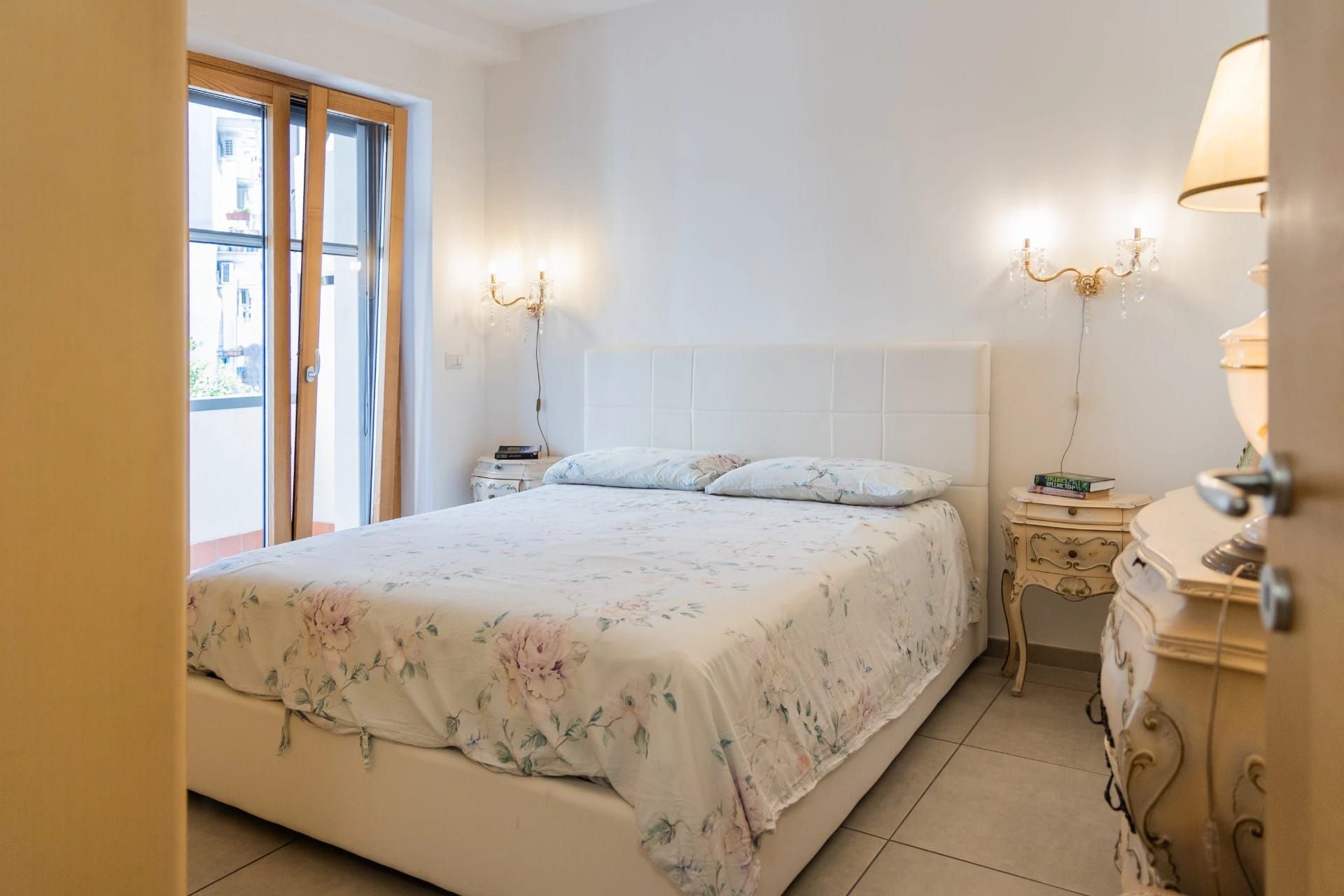 Camera da letto luminosa con letto bianco, biancheria da letto a motivi floreali, applique a parete e un'ampia finestra.