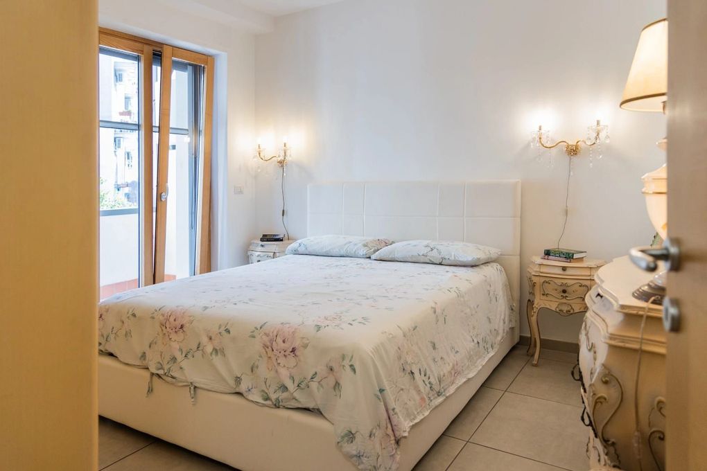 Camera da letto luminosa con copriletto floreale, pareti bianche e finestra con tende leggere.