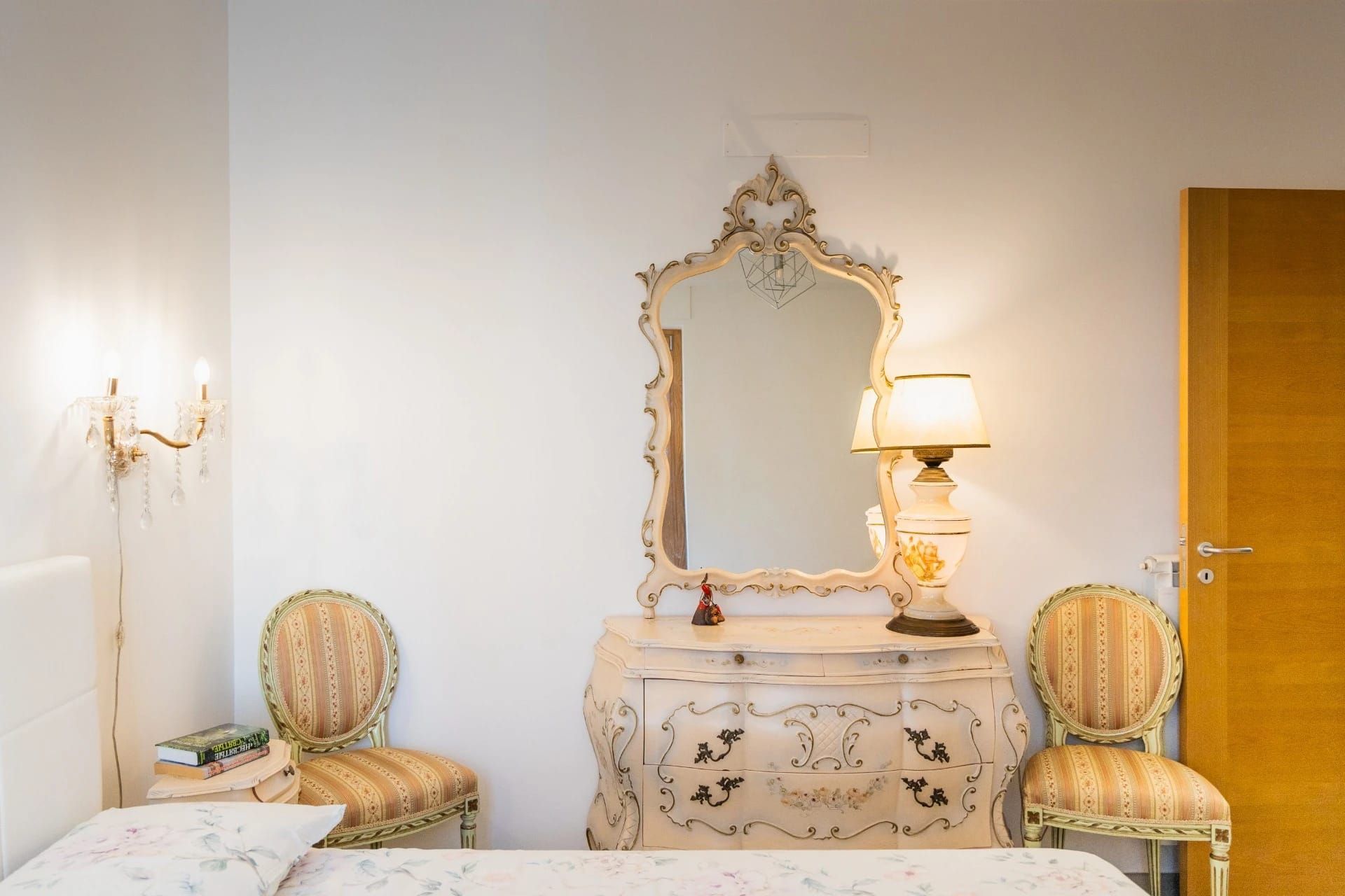 Elegante camera da letto con comò decorato, specchio, lampada e due poltrone imbottite su pareti bianche.