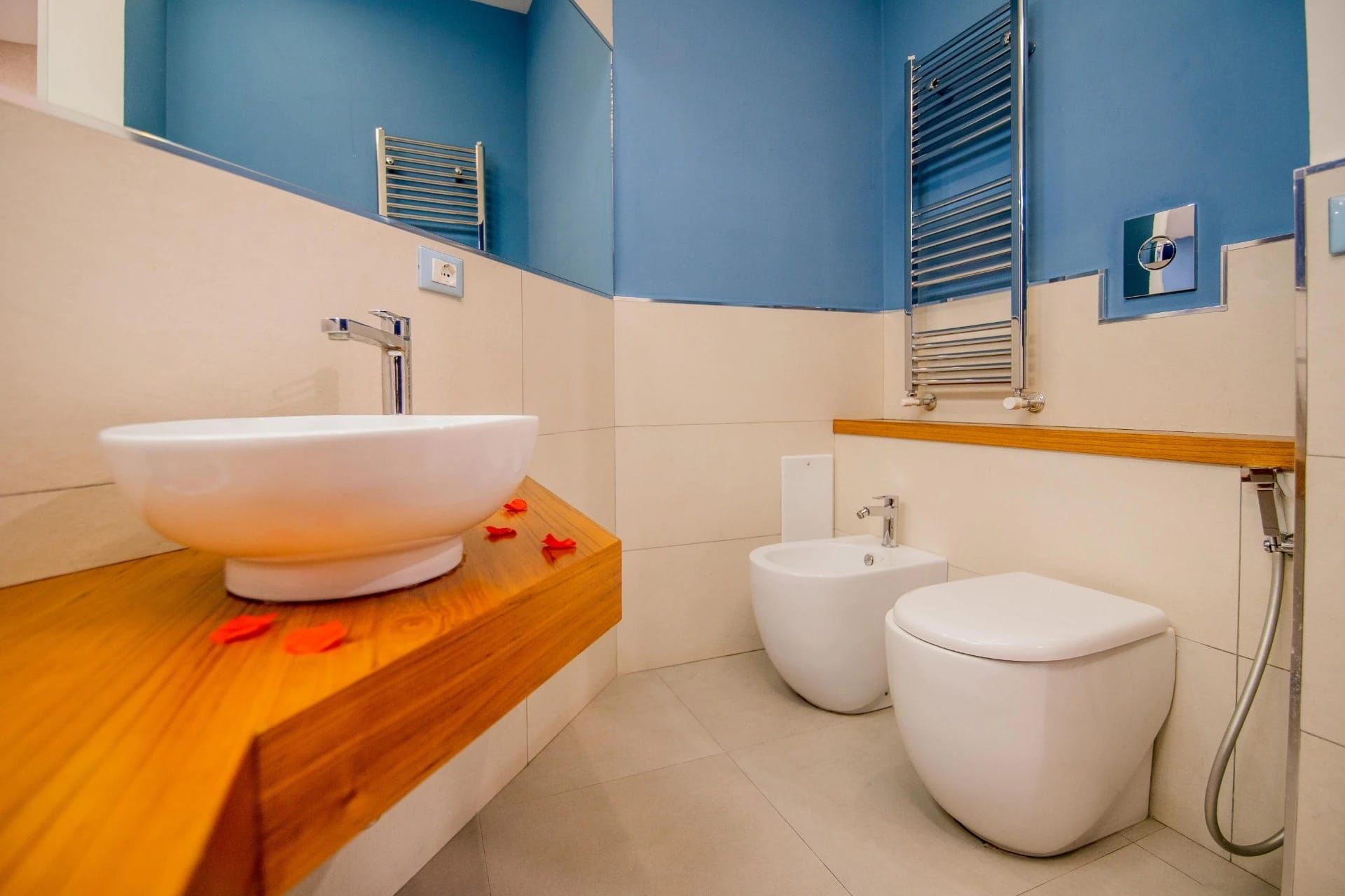 Bagno moderno con pareti blu, piano lavabo in legno e due WC bianchi accanto a un bidet.