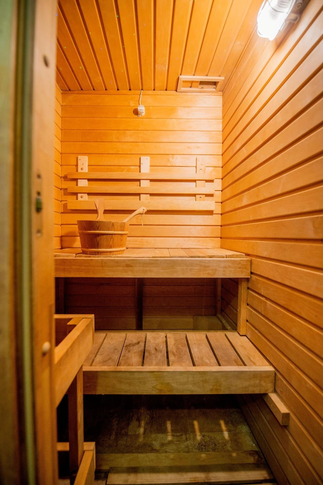 Sauna rivestita in legno con panche in legno e stufa, illuminata da una lampada a soffitto.
