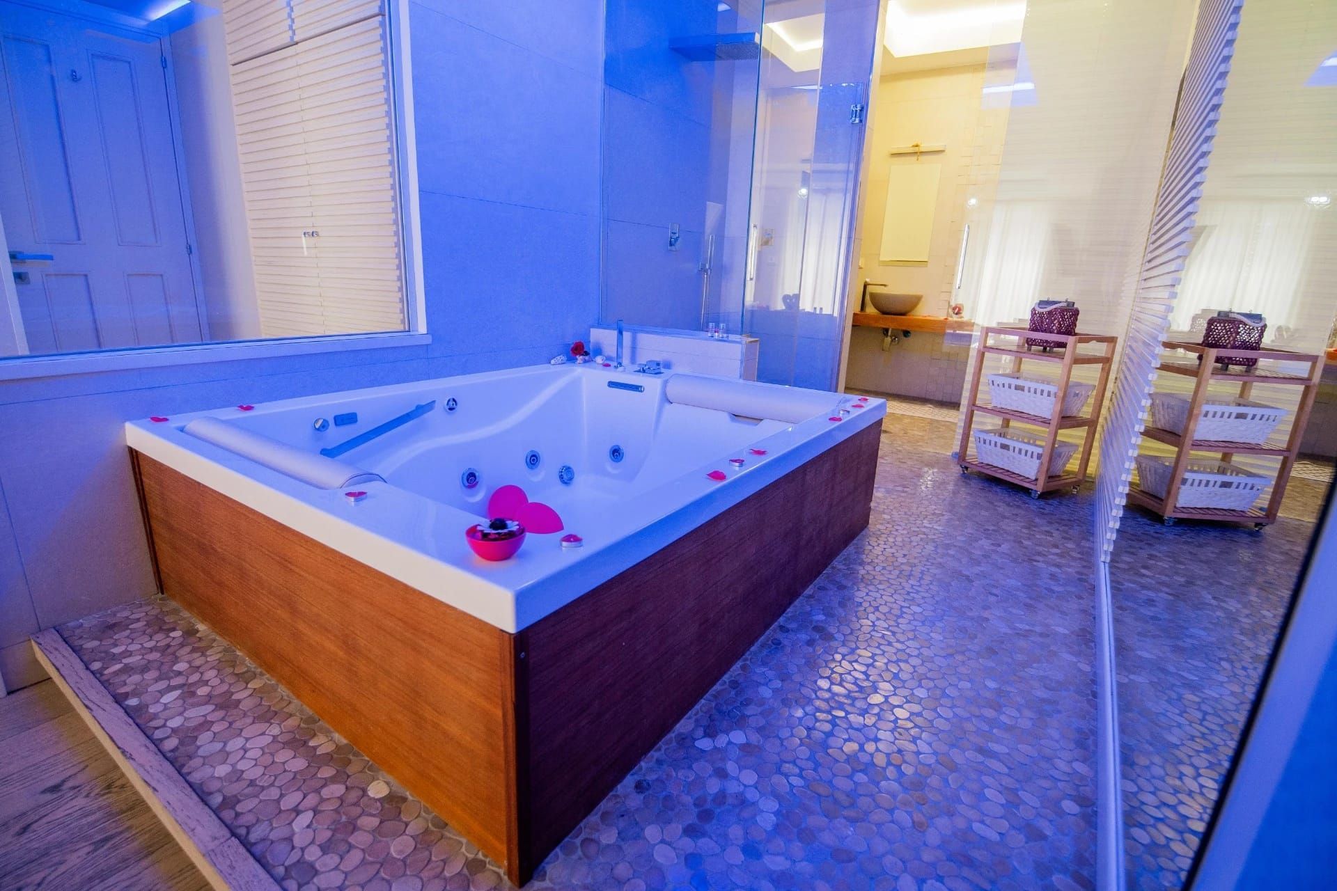 Vasca idromassaggio interna con pannelli in legno, illuminazione blu e due sedie in vimini in una sala spa piastrellata.