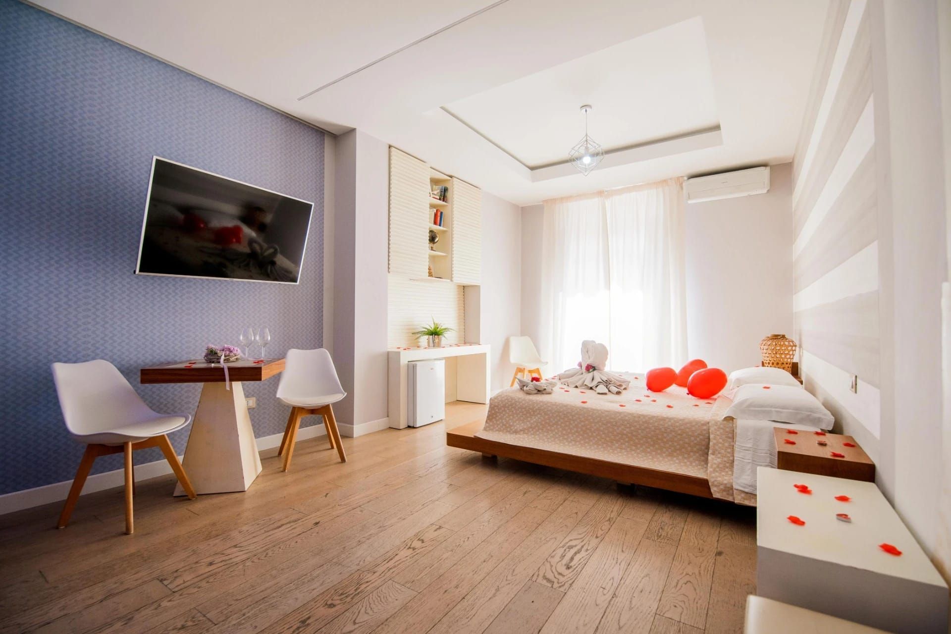 Luminosa camera da letto moderna con letto, scrivania, TV a parete e ampia finestra con tende trasparenti.