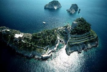 Veduta aerea di una lussureggiante isola verde con scogliere a terrazze, circondata da acque di un blu intenso e da piccoli isolotti rocciosi.