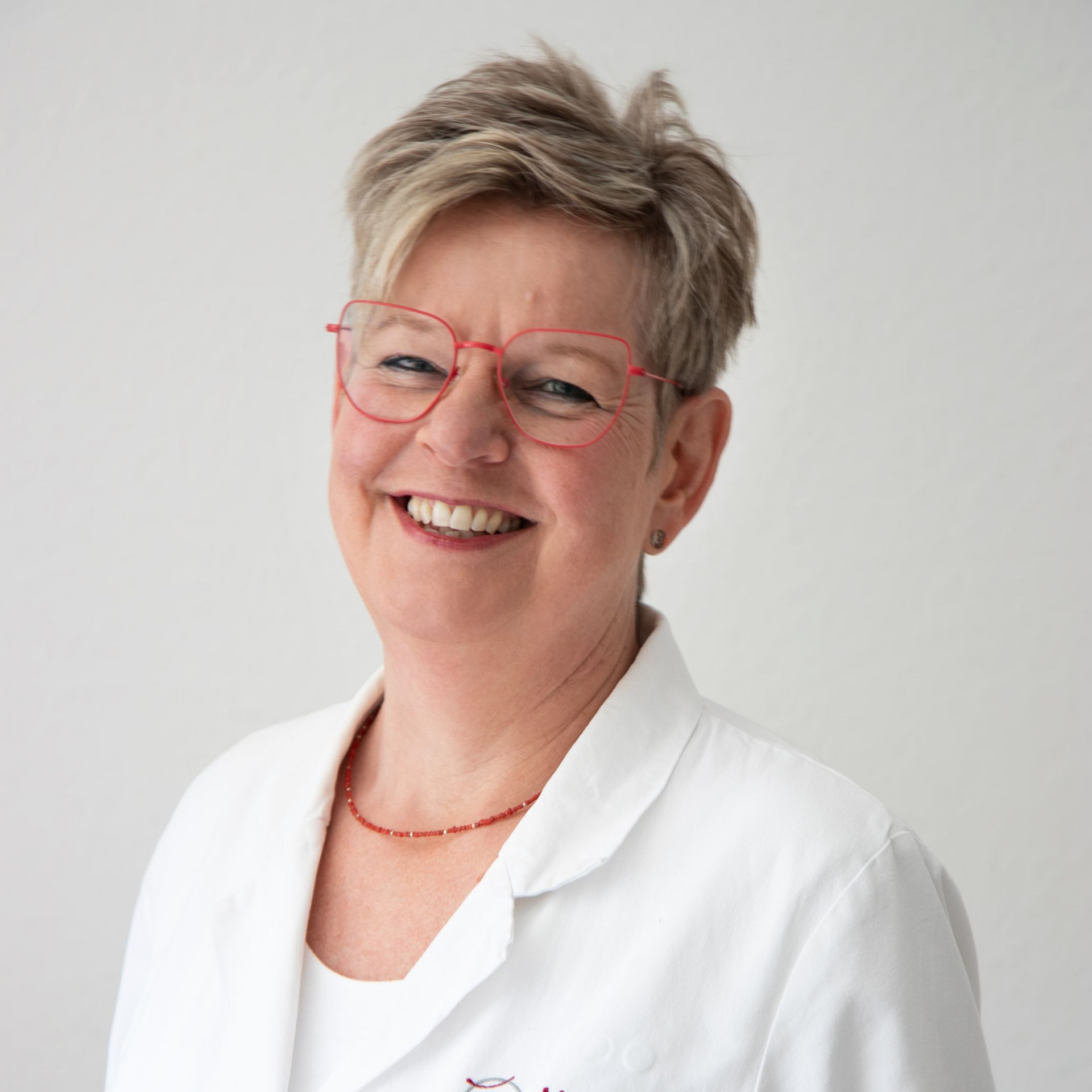 Eine Frau mit roter Brille und weißem Laborkittel lächelt.