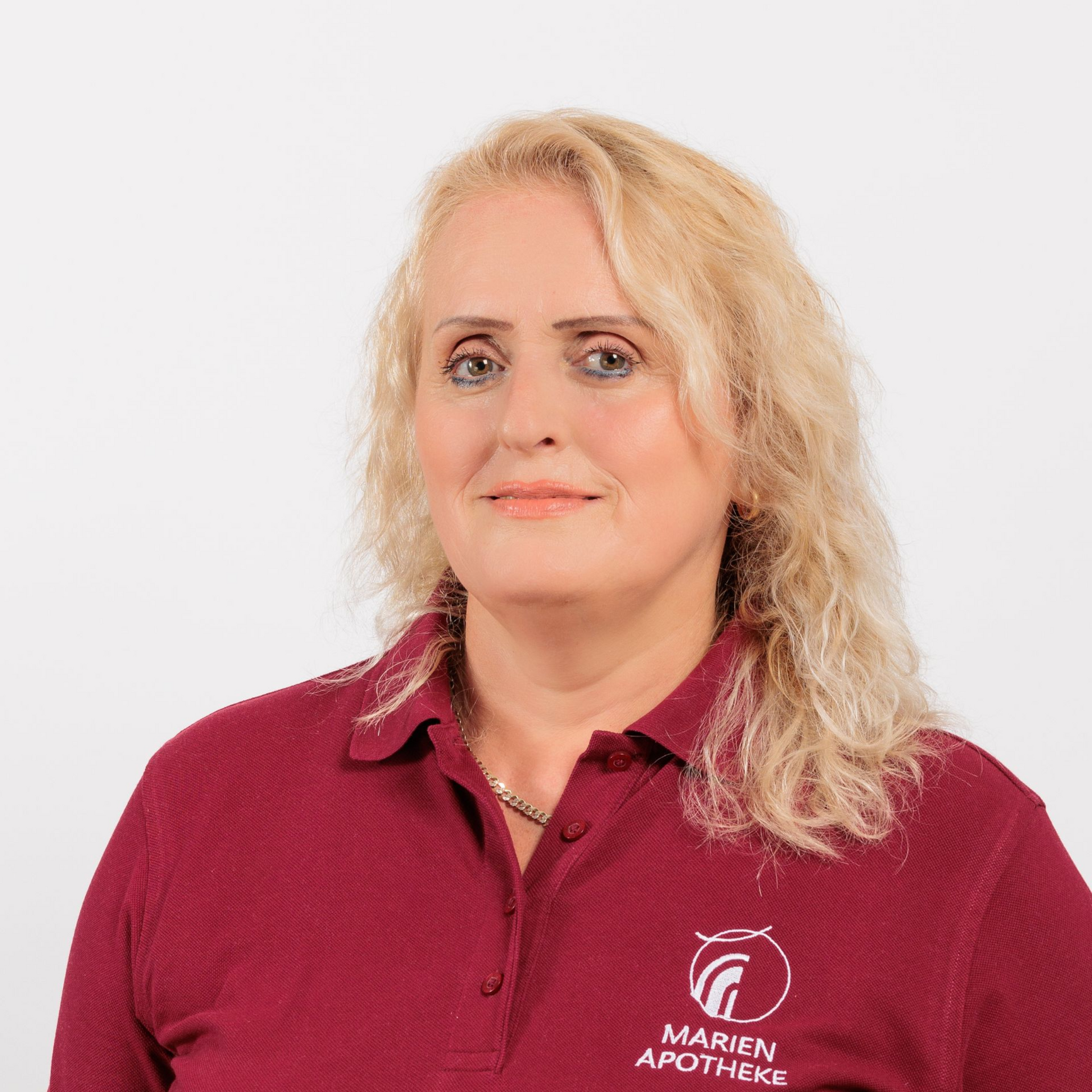 Frau mit blonden Haaren, die ein weinrotes Hemd mit Logo trägt, lächelt vor einem weißen Hintergrund.