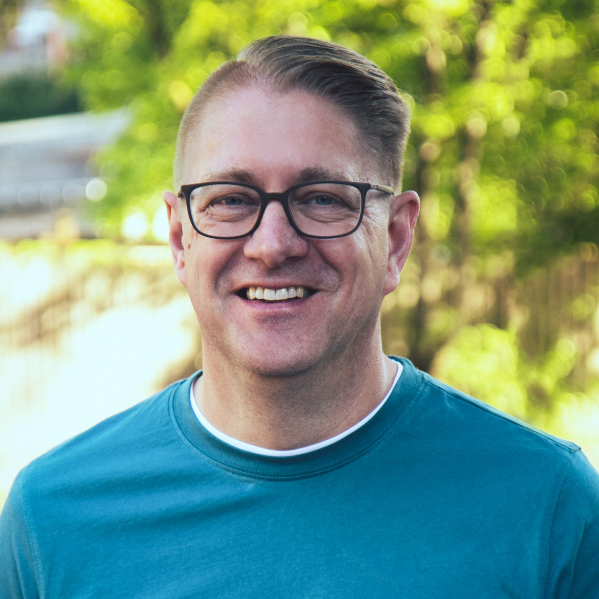 Ein Mann mit Brille lächelt, er trägt ein blaues Hemd und steht im Freien vor verschwommener grüner Kulisse.