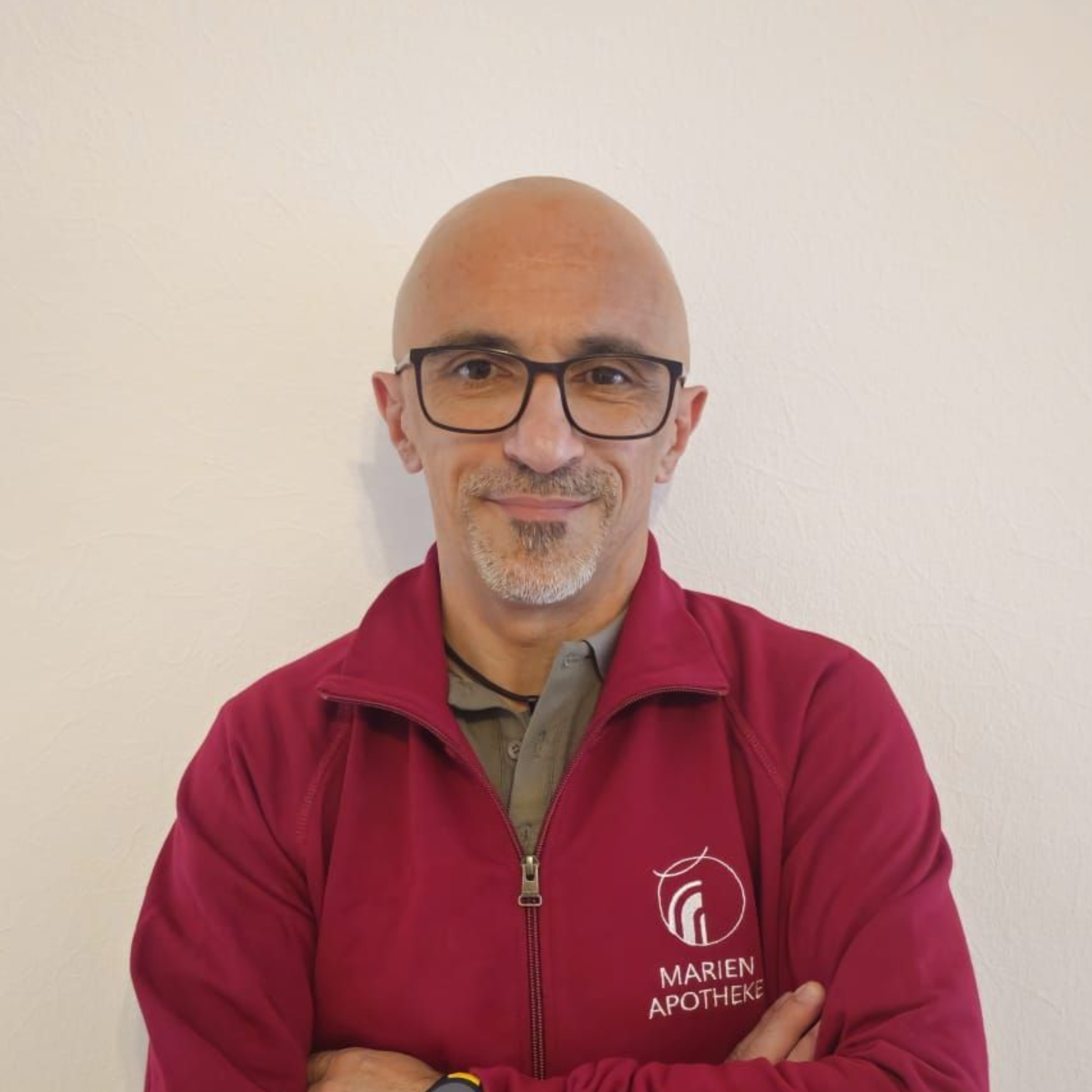 Mann mit Brille, Glatze und bordeauxroter Jacke mit Logo, die Arme verschränkt, lächelt vor einer weißen Wand.