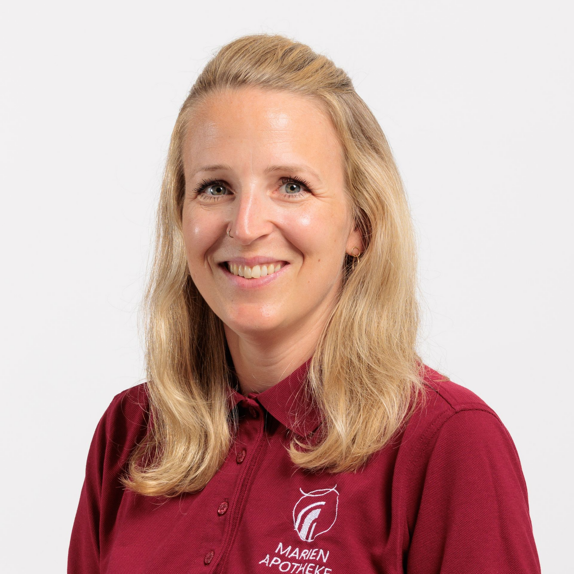 Eine Frau in einem bordeauxroten Poloshirt mit Apothekenlogo lächelt und schaut den Betrachter an.