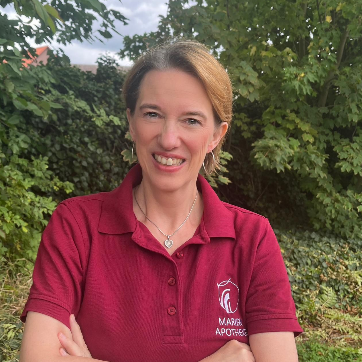 Frau mit verschränkten Armen, lächelnd, trägt ein weinrotes Poloshirt im Freien.