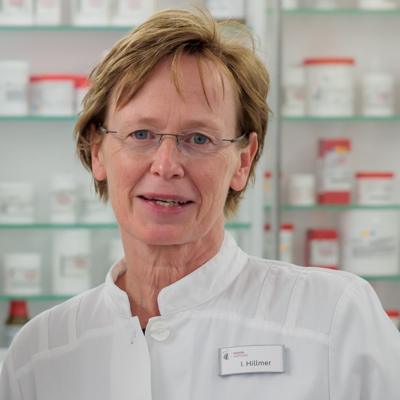 Frau im weißen Kittel und mit Brille lächelt in einer Apotheke; im Hintergrund sind Regale mit Medikamenten zu sehen.