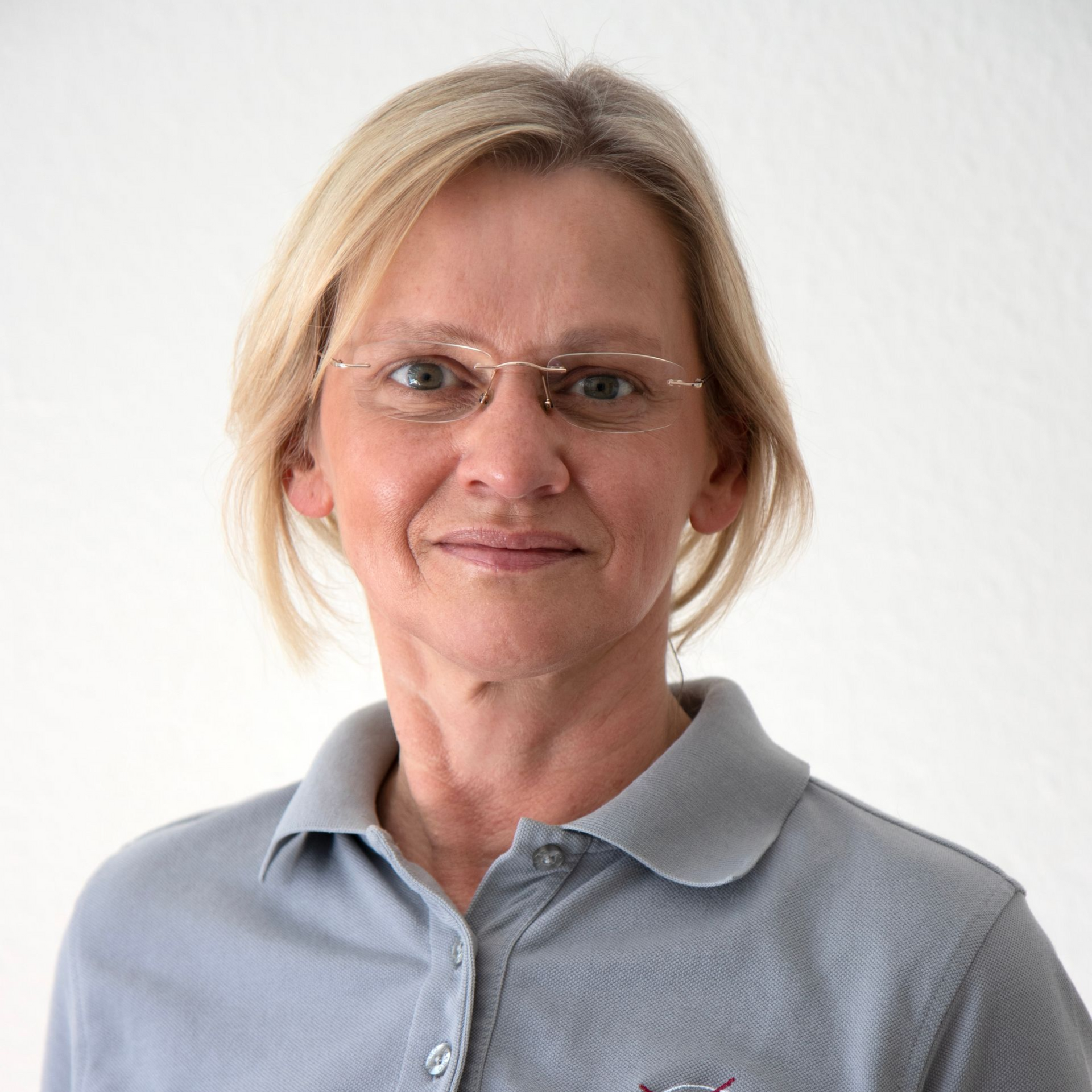 Frau mit Brille lächelt, trägt ein graues Poloshirt, vor weißem Hintergrund.