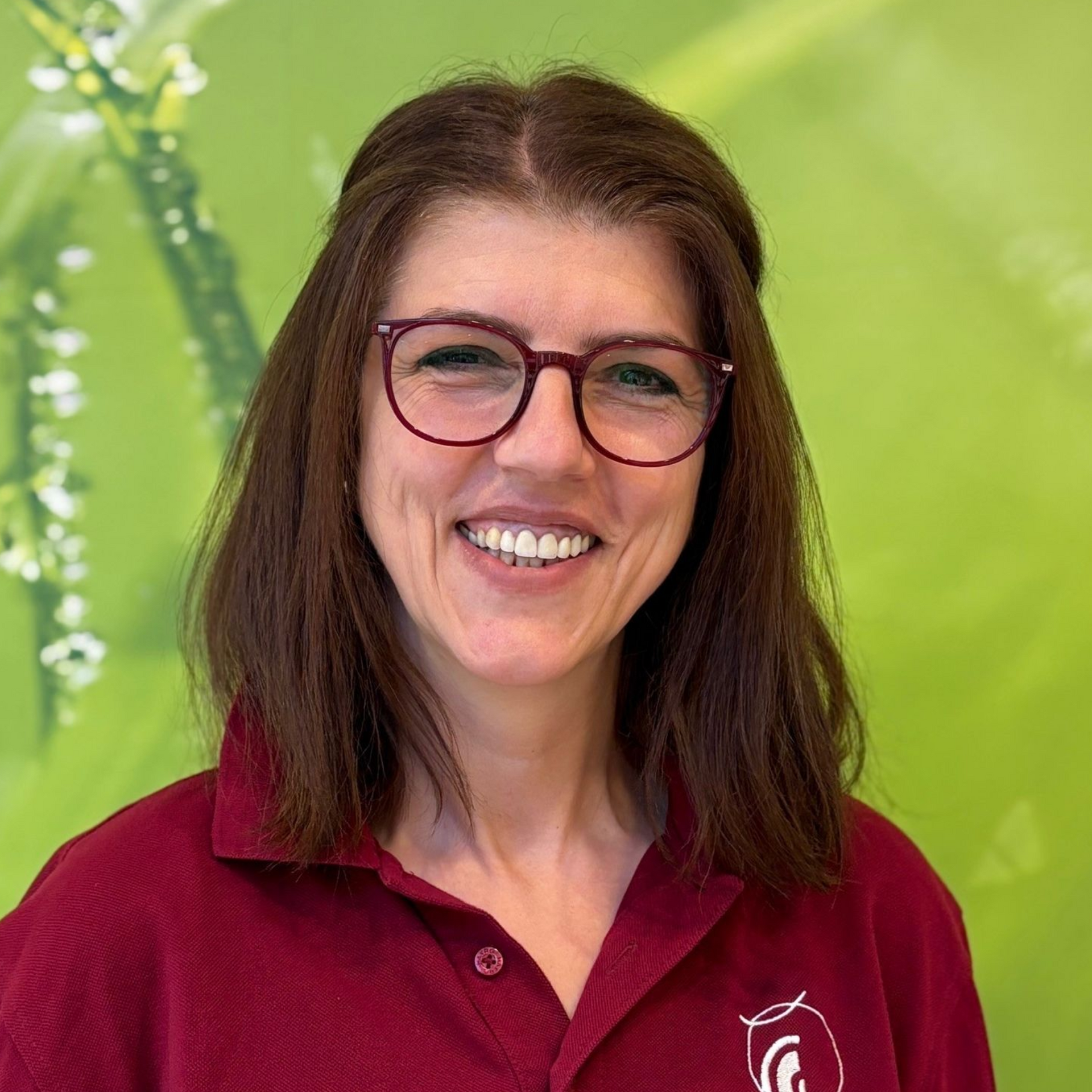 Frau mit Brille lächelt, sie trägt ein bordeauxrotes Poloshirt vor grünem Hintergrund.
