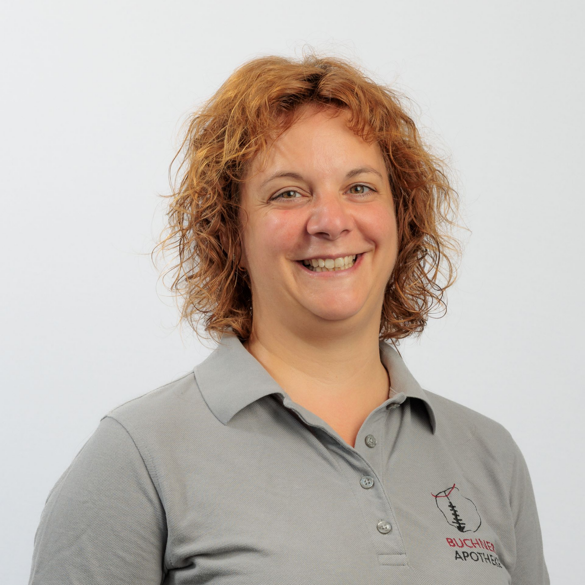 Eine Frau mit lockigem, kastanienbraunem Haar lächelt vor einem weißen Hintergrund. Sie trägt ein graues Poloshirt mit Logo.