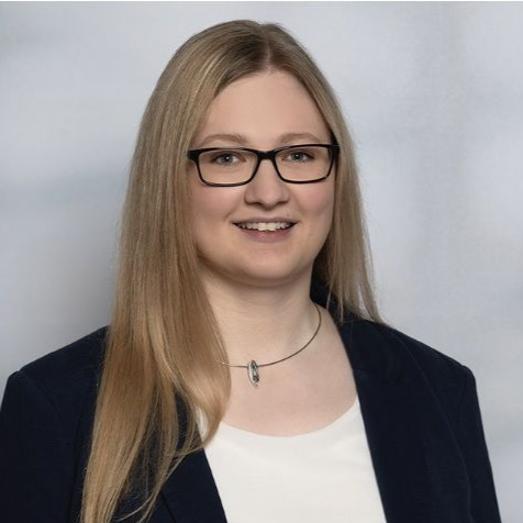 Frau mit langen blonden Haaren, Brille, die einen dunkelblauen Blazer und ein weißes Hemd trägt, lächelt.