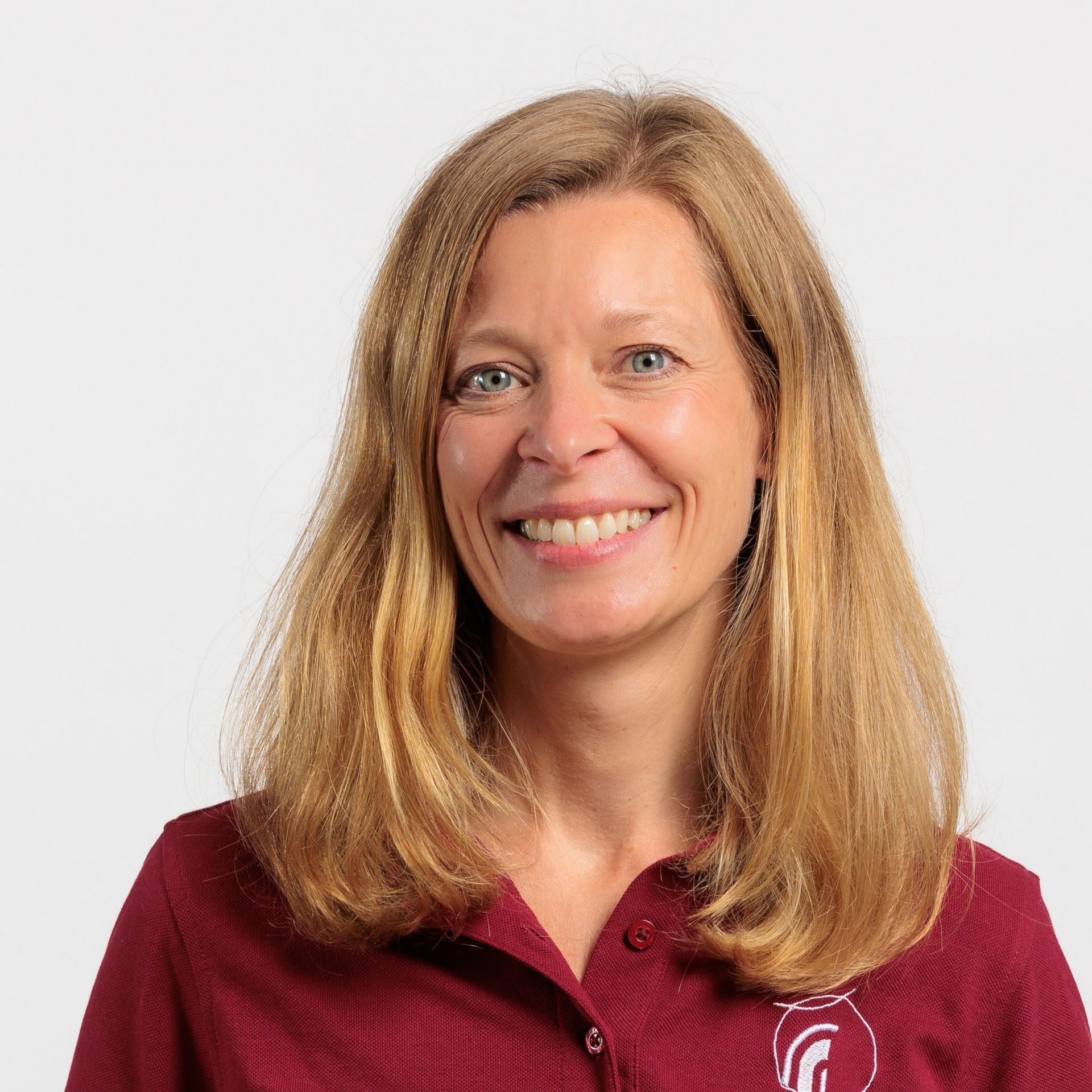 Eine Frau mit blonden Haaren, die ein weinrotes Poloshirt trägt, lächelt in die Kamera.