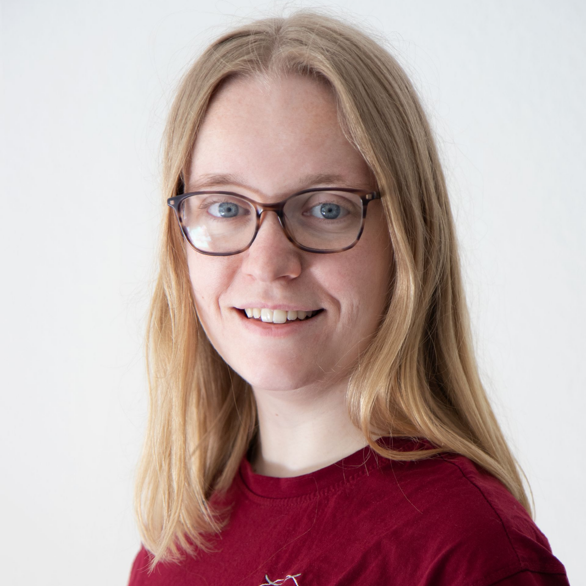 Eine Frau mit blonden Haaren und Brille lächelt vor einem weißen Hintergrund und trägt ein rotes Hemd.