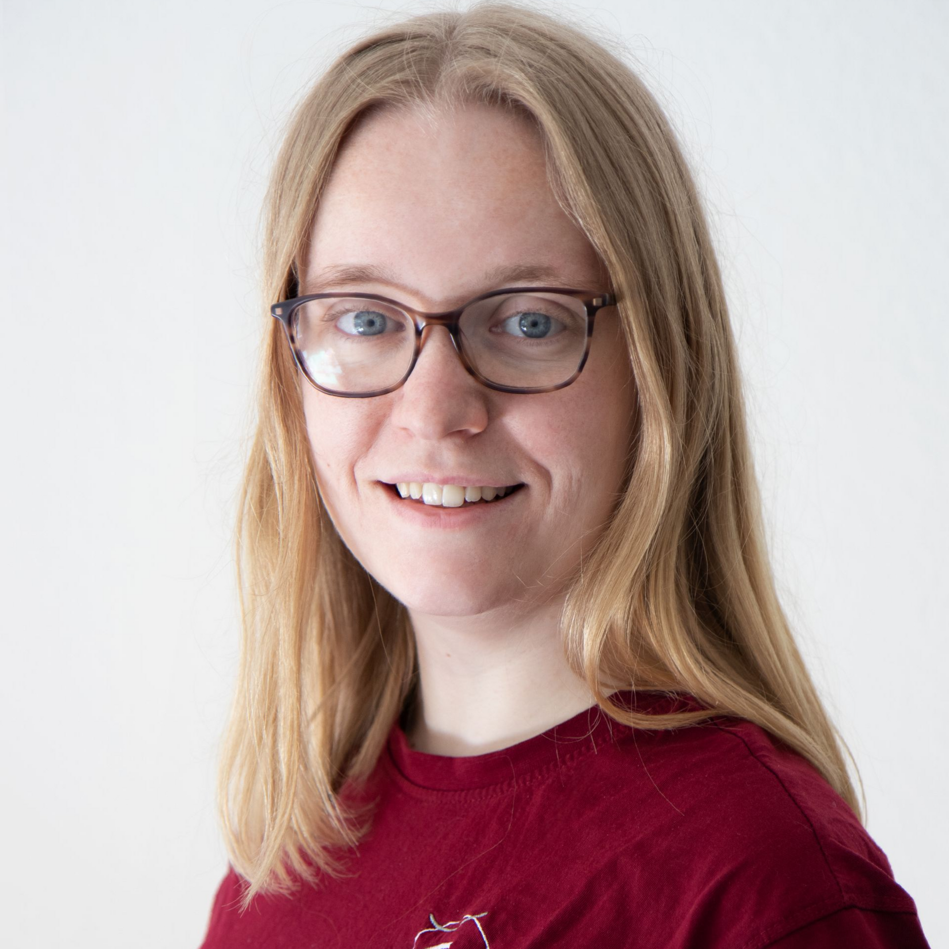 Eine Frau mit blonden Haaren und Brille lächelt in einem roten Hemd vor einem weißen Hintergrund.