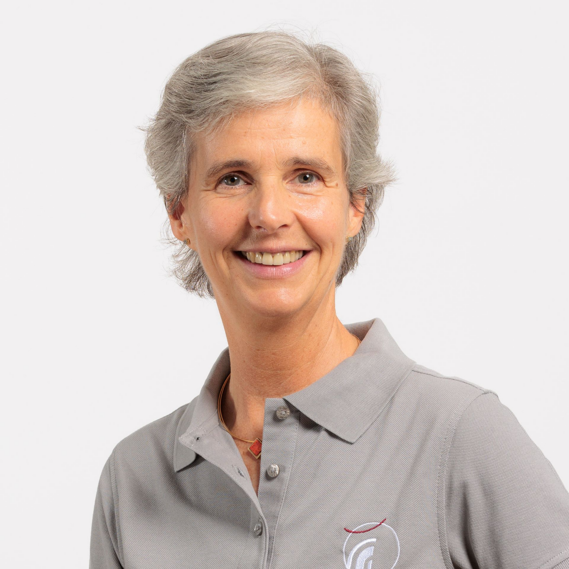 Eine Frau mit kurzen grauen Haaren lächelt in einem grauen Poloshirt vor weißem Hintergrund.