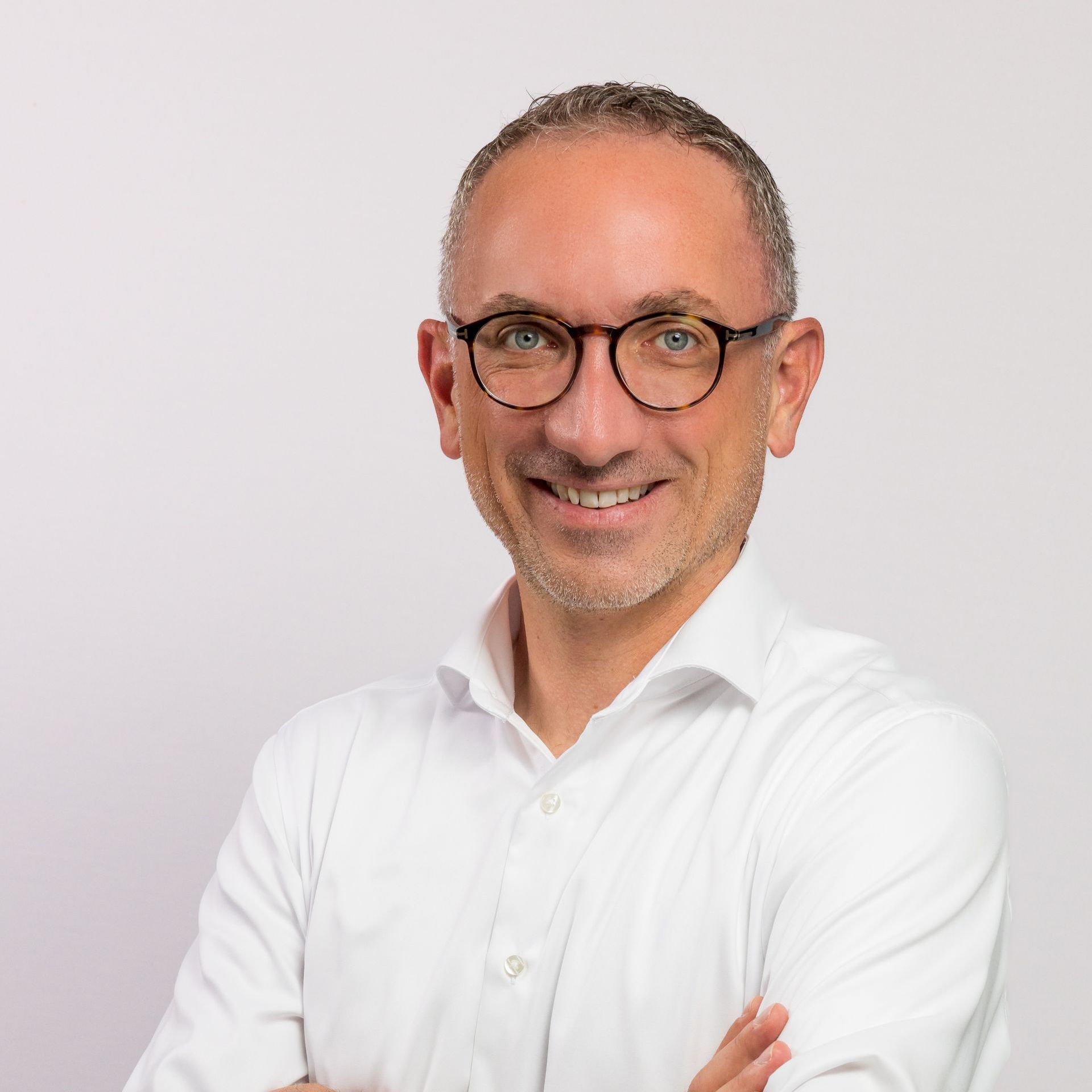 Mann mit Brille, verschränkten Armen, lächelnd, trägt ein weißes Hemd vor weißem Hintergrund.