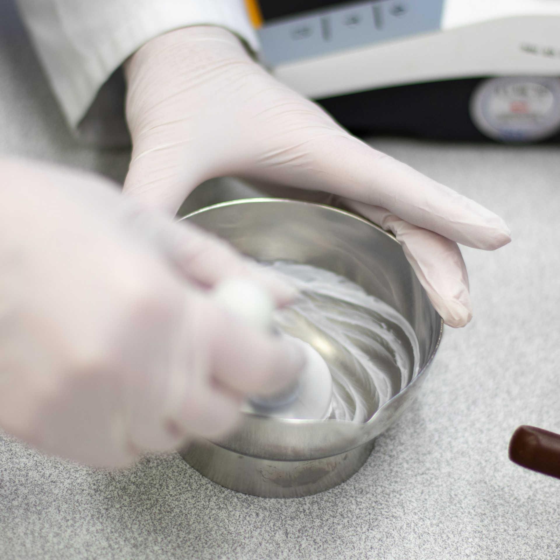 Hände in weißen Handschuhen vermischen eine Substanz in einer Metallschüssel.