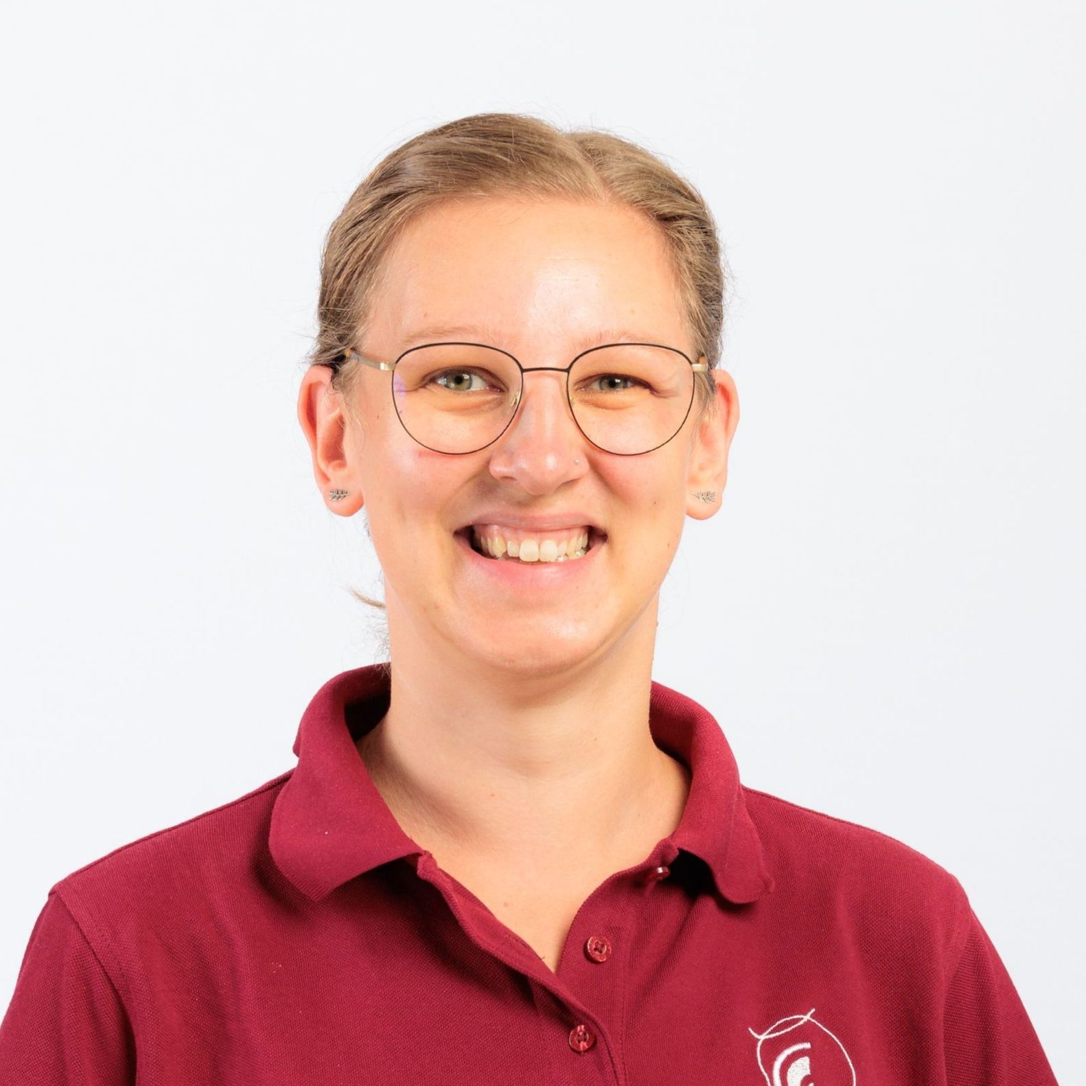 Frau mit Brille lächelt, trägt ein weinrotes Poloshirt und steht vor weißem Hintergrund.