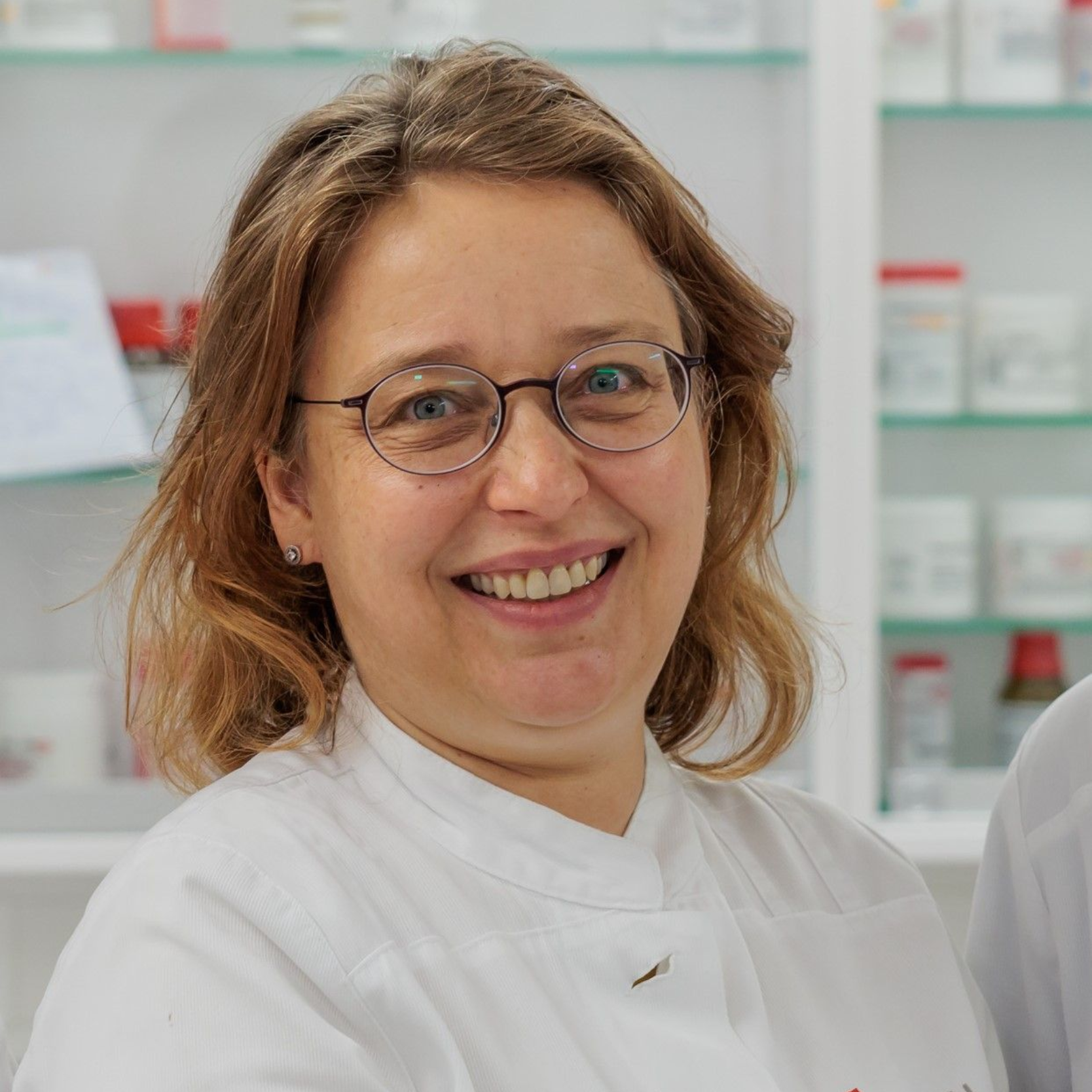 Eine Frau im weißen Kittel lächelt und trägt eine Brille. Sie befindet sich in einer Apotheke mit Regalen voller Medikamente.