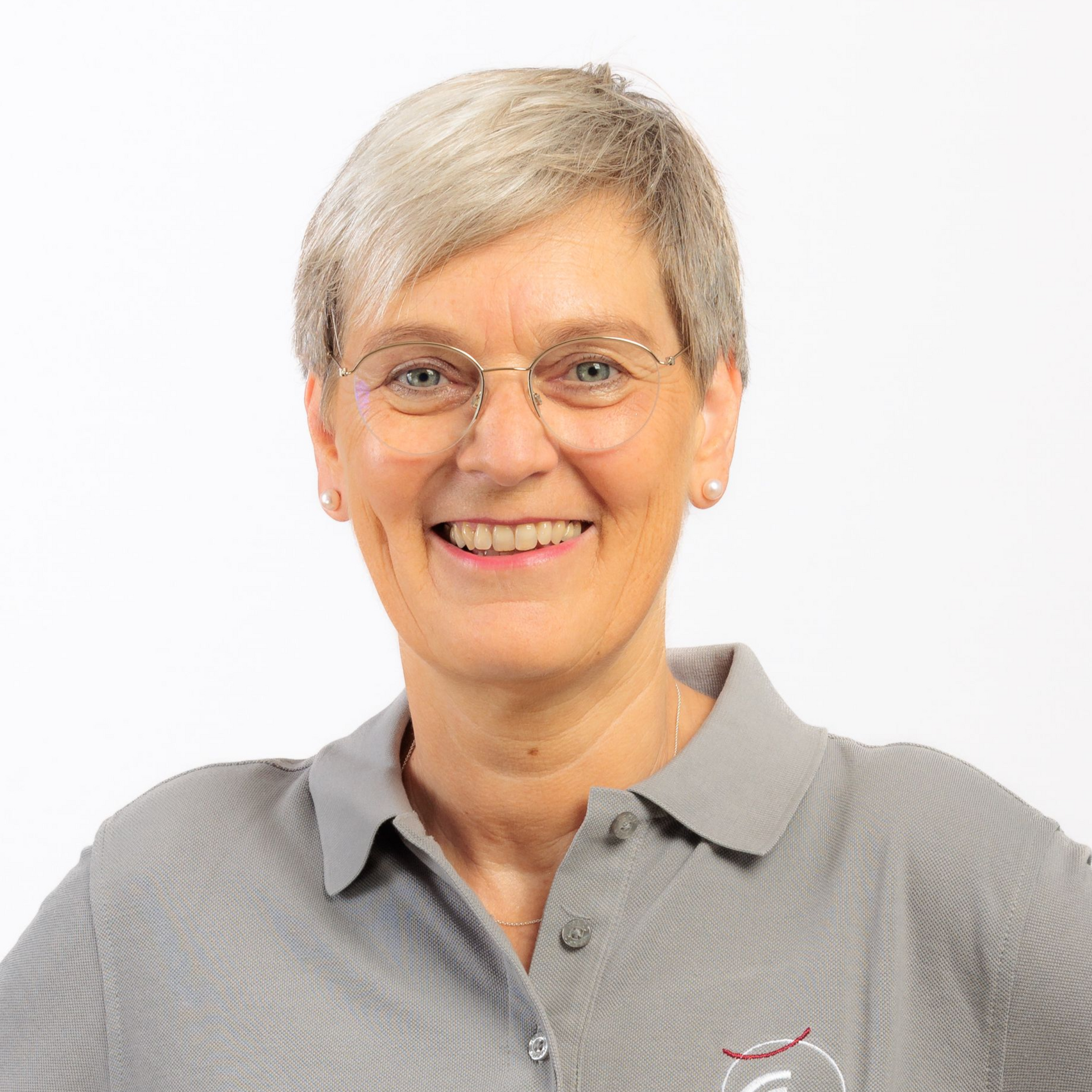Frau mit kurzem grauen Haar, Brille und grauem Poloshirt, lächelt.