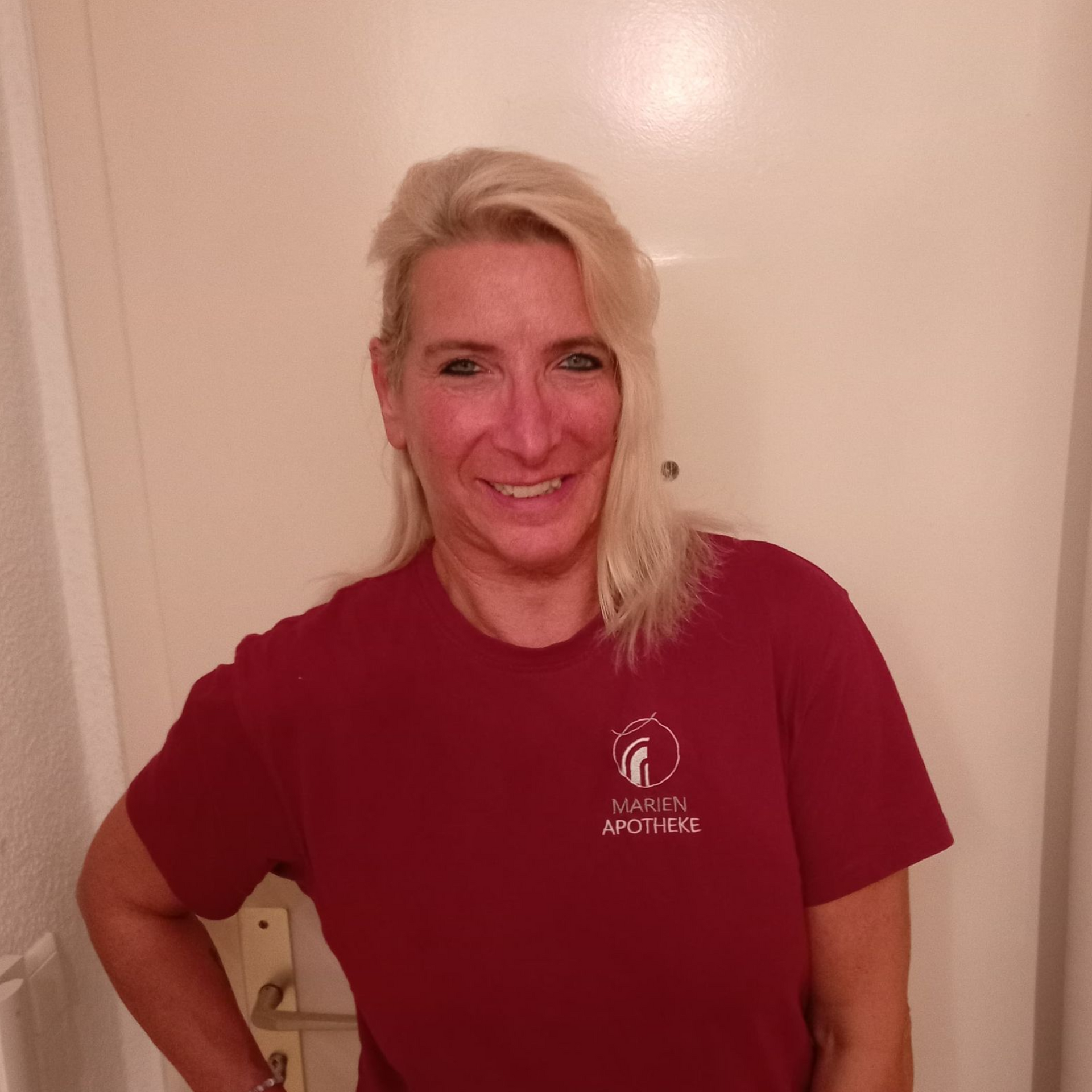 Eine Frau mit blonden Haaren und einem weinroten Hemd lächelt vor einer weißen Tür.