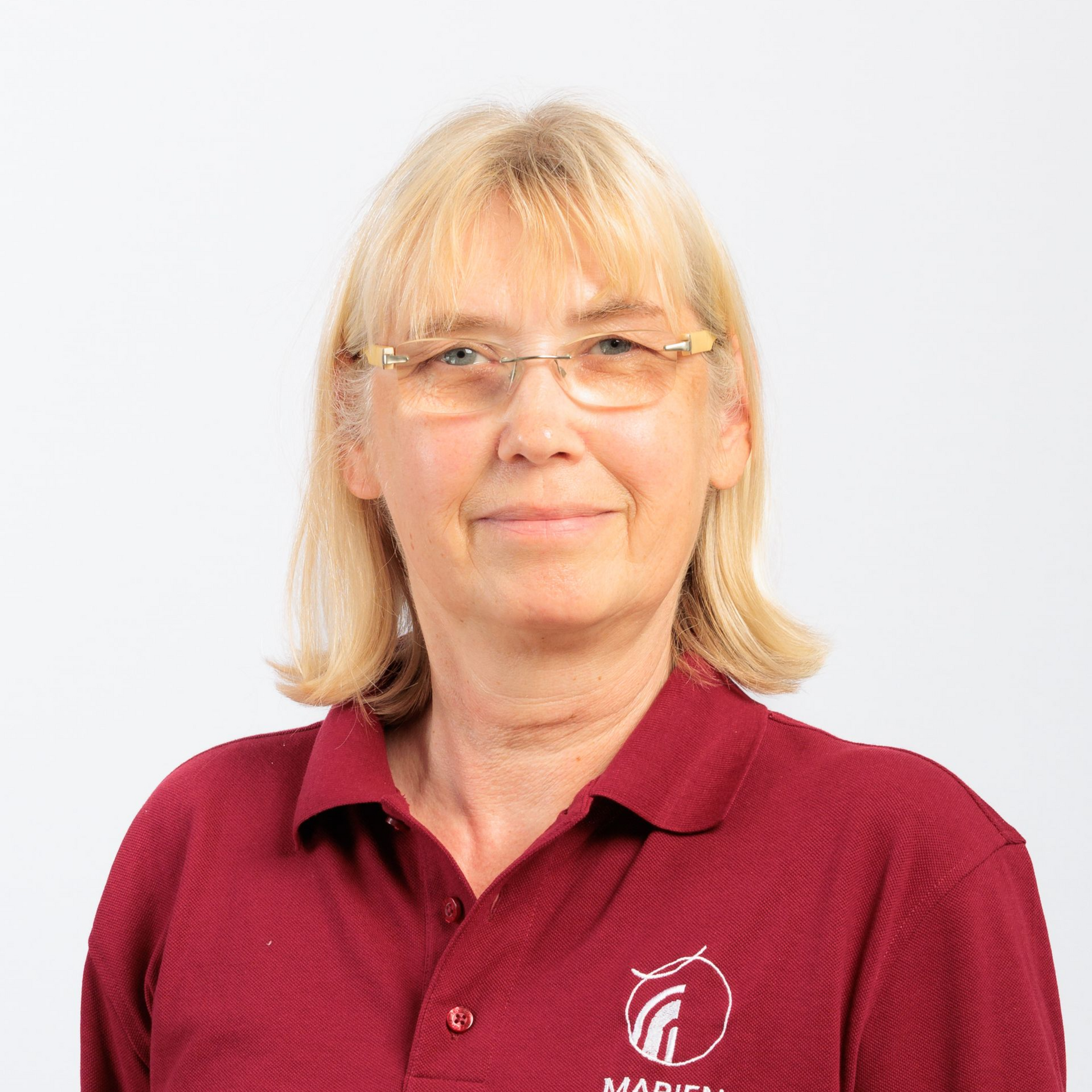 Frau mit blonden Haaren und Brille, die ein rotes Poloshirt trägt und leicht lächelt.