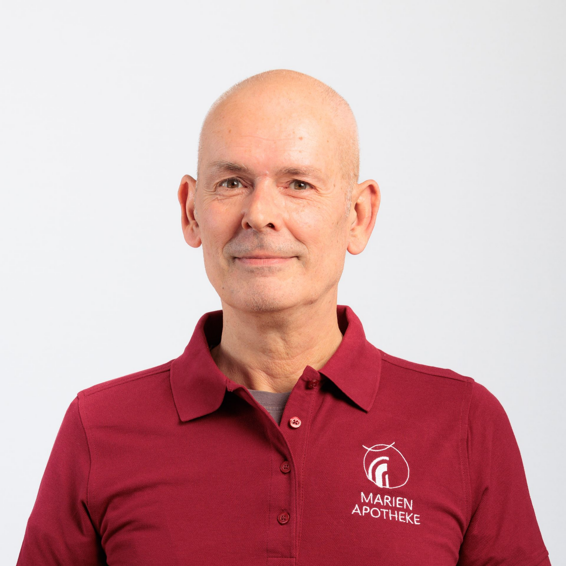 Ein Mann mit Glatze, der ein weinrotes Poloshirt mit Logo trägt, vor einem weißen Hintergrund.